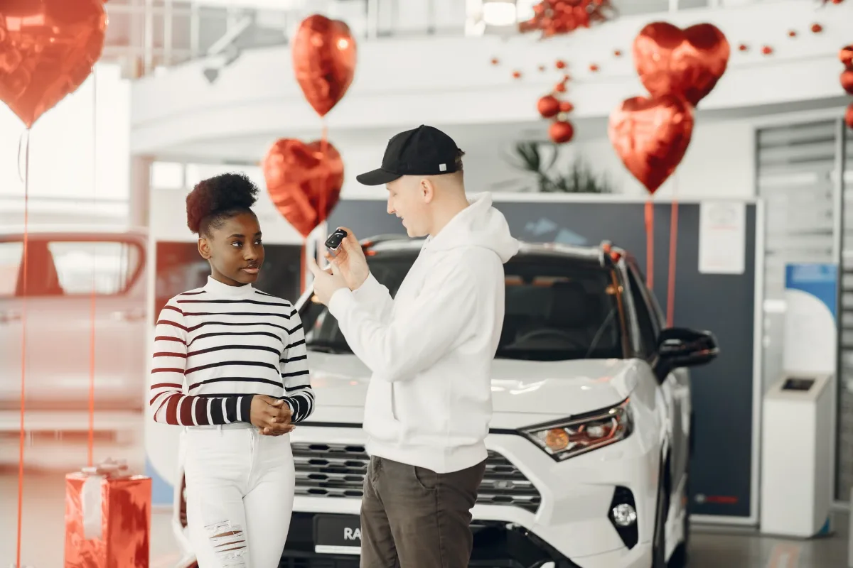 A couple exchanging car keys inside a showroom decorated with heart-shaped balloons, celebrating a new car purchase